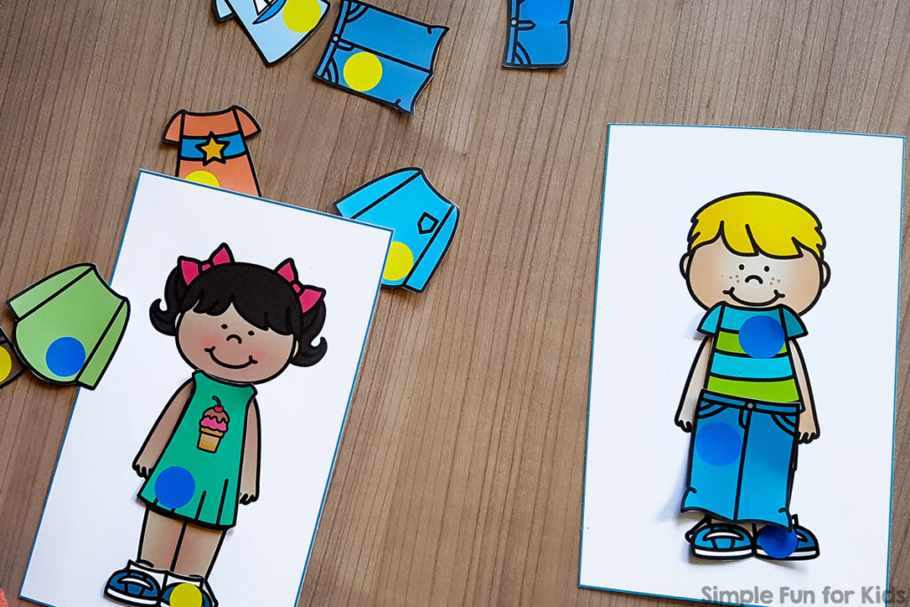 Two paper dolls of a boy and girl. The girl is wearing a turquoise dress with a blue circle on it and blue shoes with a yellow dot on them. The boy is wearing a blue and green shirt with a blue dot, blue pants with a blue dot, and blue shoes with a blue dot on them.