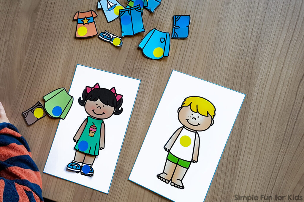 A boy paper doll with a yellow circle on the chest. Next to it, there's a girl paper doll wearing a dress with an ice cream cone on the front and a blue circle. On the girl paper doll's feet are blue shoes with a blue dot on them. Other items of paper clothing are scattered around: shorts, shirts, shoes, pants, etc.