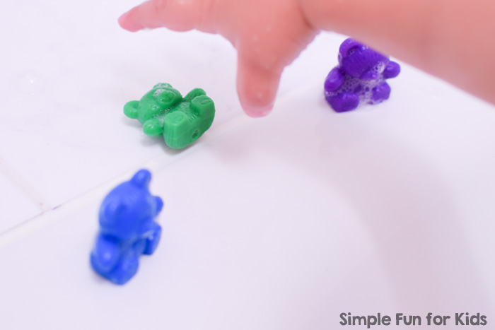 Toddler playing with a blue, green, and purple rainbow bear in the bathtub.