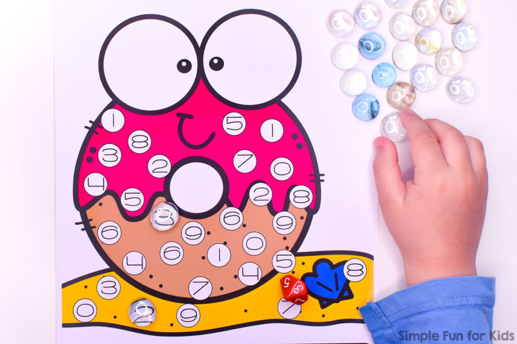 Kindergarten-aged child playing a roll and cover game to practice number recognition. The gameboard features a cute beach donut image with large eyes and numbers 0-9. He uses a 10-sided dice and glass gems as manipulatives to cover the numbers he rolls with the dice.