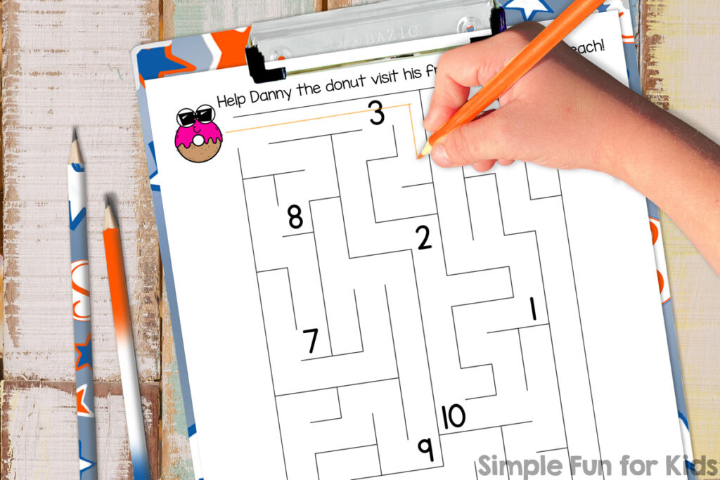 Image of summer donuts number maze on a clipboard on top of a wooden desk. Next to the printable, there are two pencils. Also shown is a kindergarteners hand holding an orange pencil and following the path of the maze.