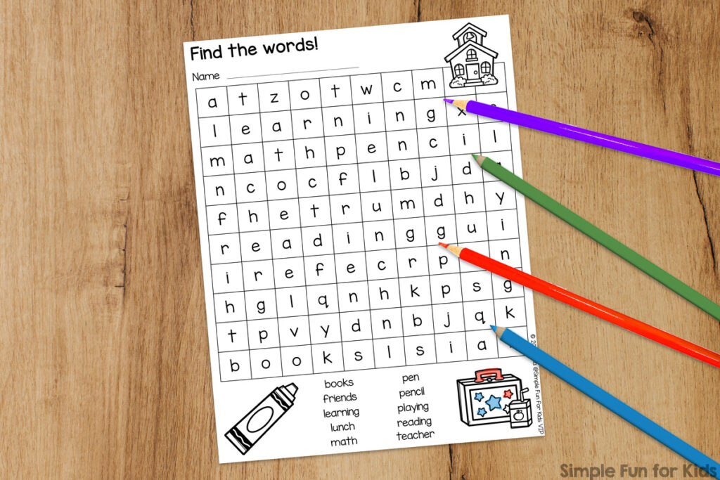 A picture of the black & white version of the back to school word search that's partially colored in. It's on top of a wooden desktop and next to some colored pencils in blue, red, and green.