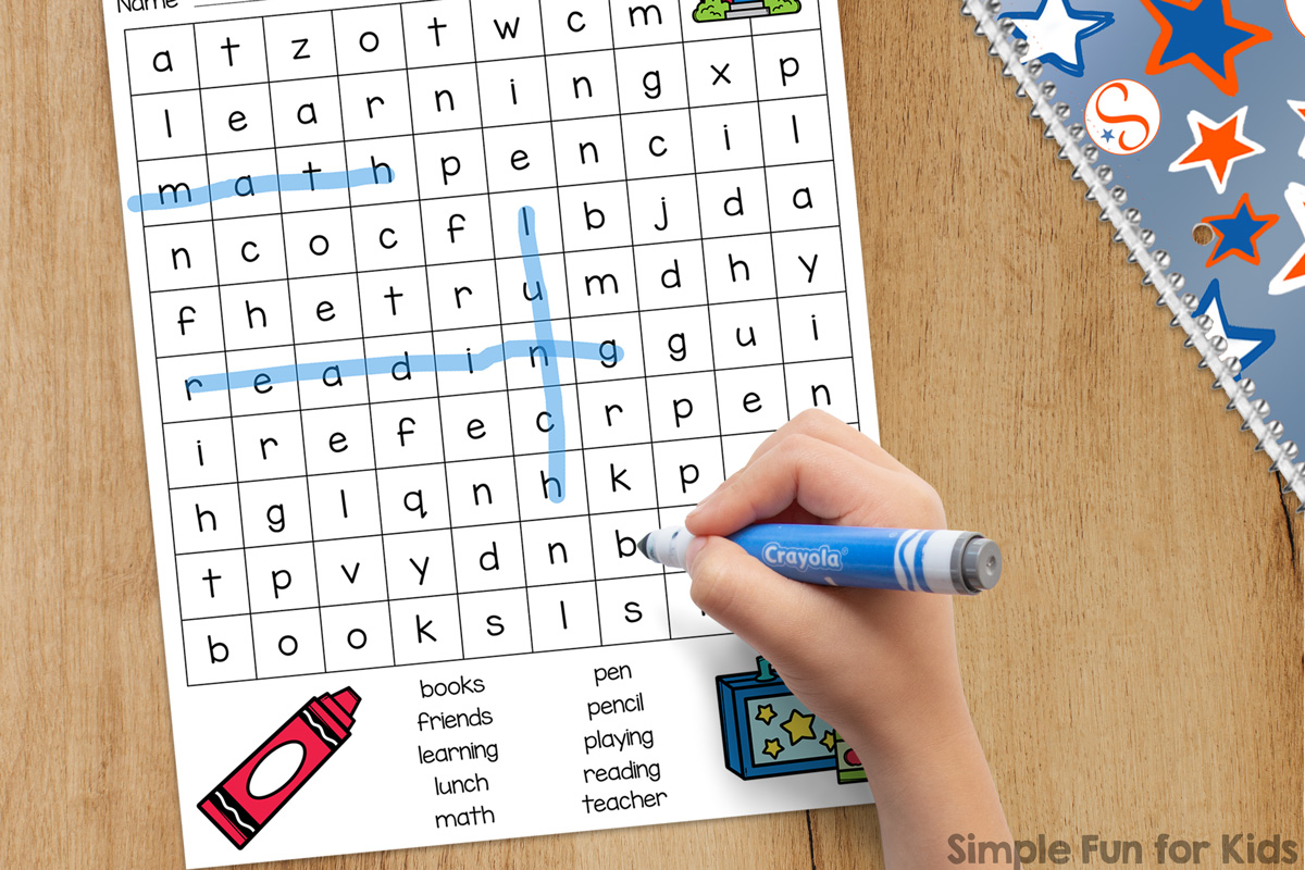 Picture of the one-page Back to School Word Search on top of a light wooden desk next to a Simple Fun for Kids notebook. A first-grade student is holding a blue marker and solving the word search puzzle.