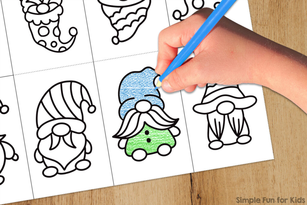 Picture of one of the Gnomes Mini Folding Coloring Books on a light brown wooden desk with a student's hand holding a light blue colored pencil while coloring a gnome's hat. The gnome's sweater is colored green. The Simple Fun for Kids watermark is at bottom right.